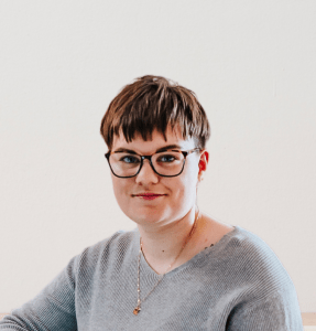 Headshot of Jen. They are a white non-binary person with blue eyes and short brown hair styled in a textured bowlcut. They wear purple glasses with yellow tortoiseshell on the top of the frames, a ribbed grey v-neck sweater, and a gold necklace.